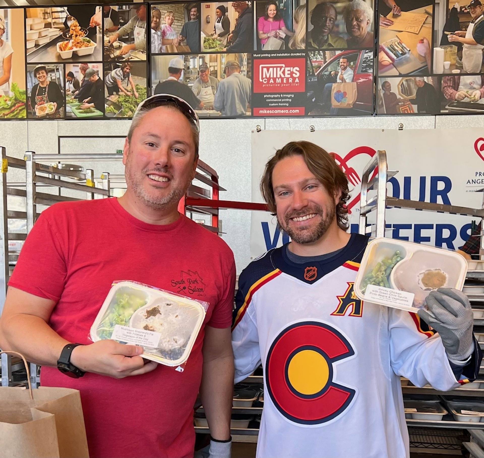 Two men in hockey jerseys proudly display a variety of food items, showcasing their enthusiasm for volunteering. .