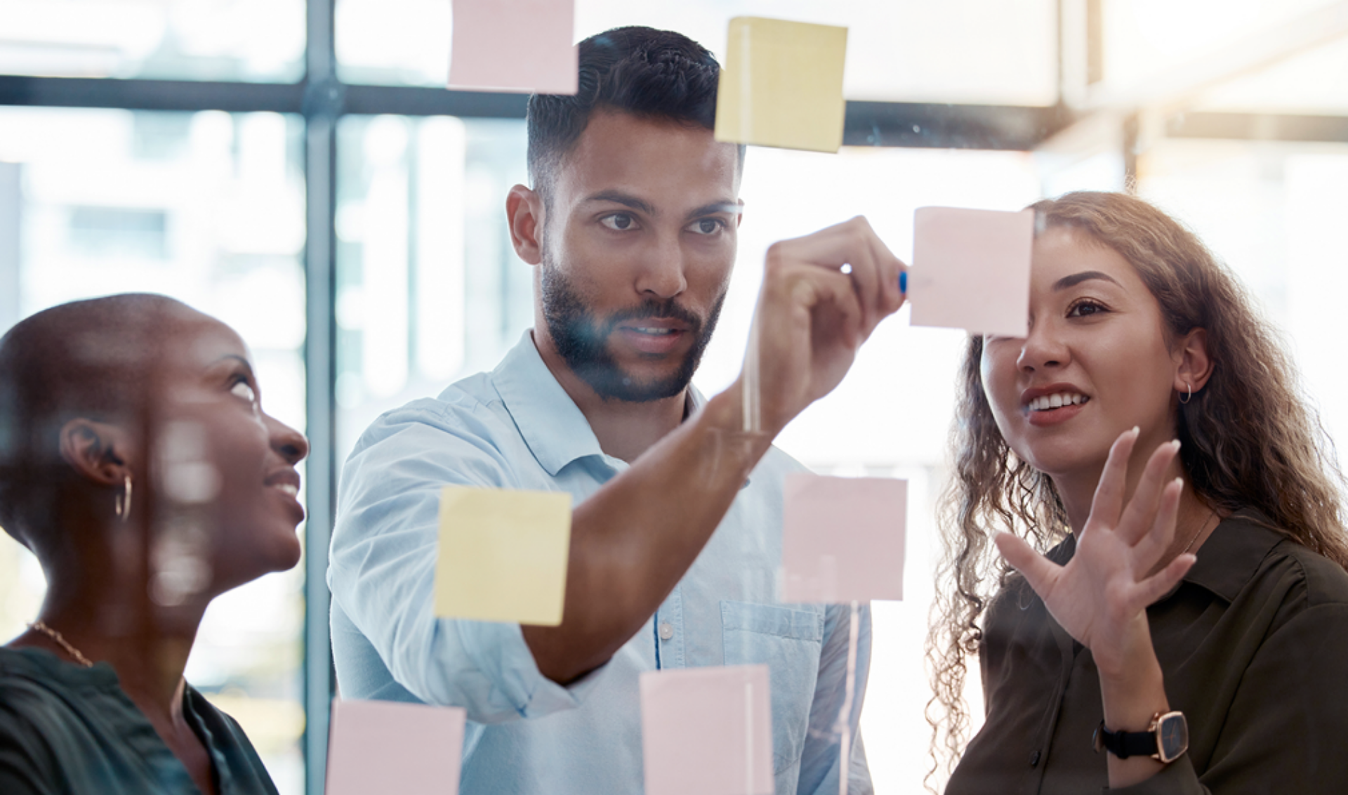 Three individuals examine colorful sticky notes affixed to a glass wall, engaged in discussion and brainstorming ideas.