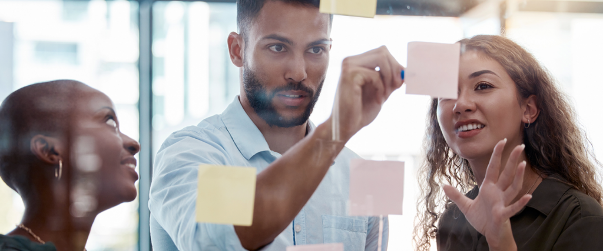 Three individuals examine colorful sticky notes affixed to a glass wall, engaged in discussion and brainstorming ideas.