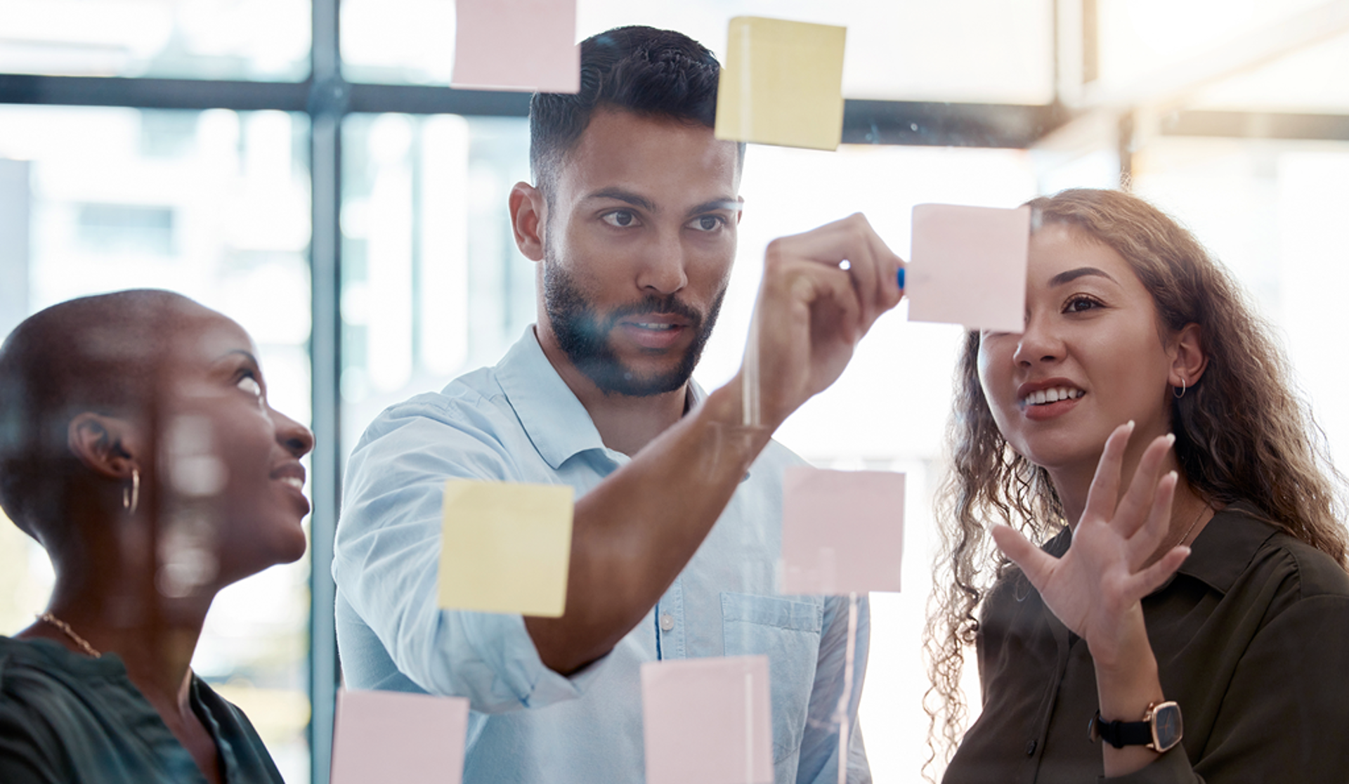 Three individuals examine colorful sticky notes affixed to a glass wall, engaged in discussion and brainstorming ideas.