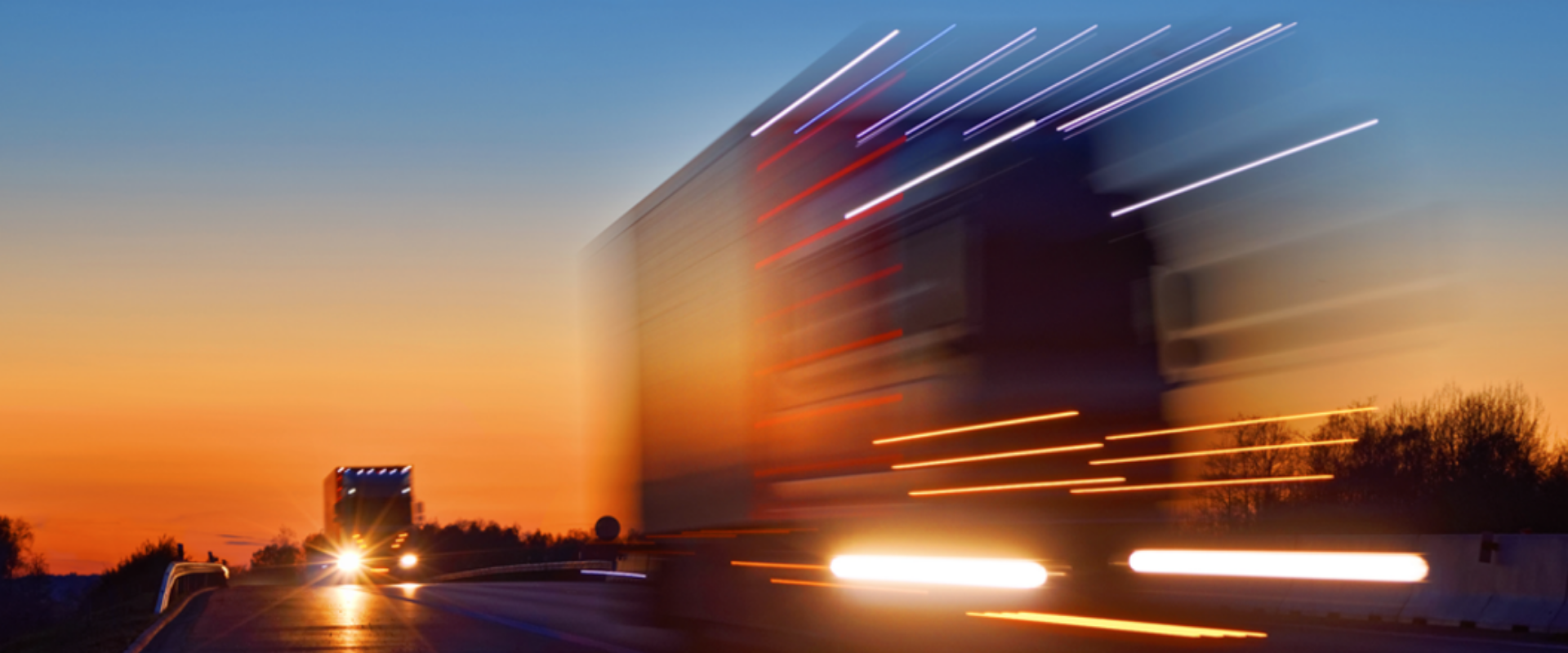 A truck moves along a highway at sunset, with the sky ablaze in shades of orange and pink, creating a picturesque scene.