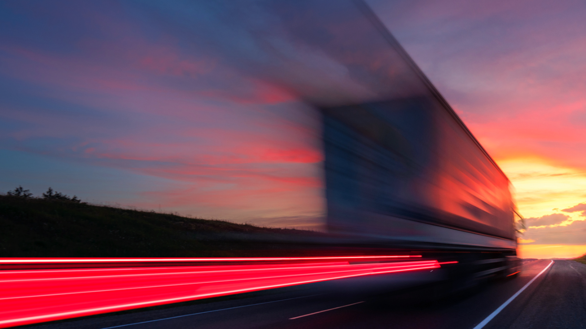 A truck moves down a highway, with a picturesque sunset casting warm hues across the scene.
