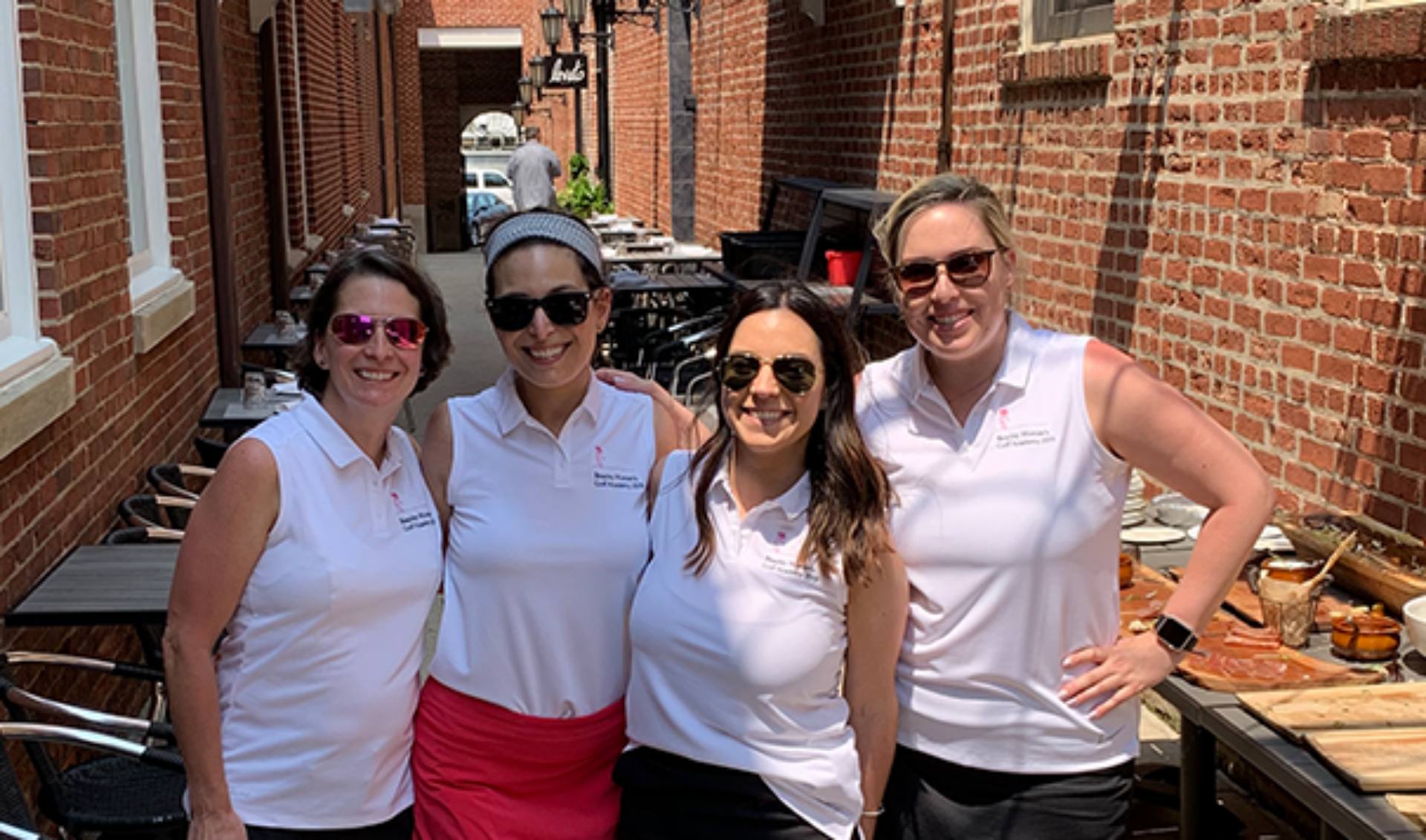 Four women dressed in white shirts and black skirts pose together for a group photo, smiling and looking confident.
