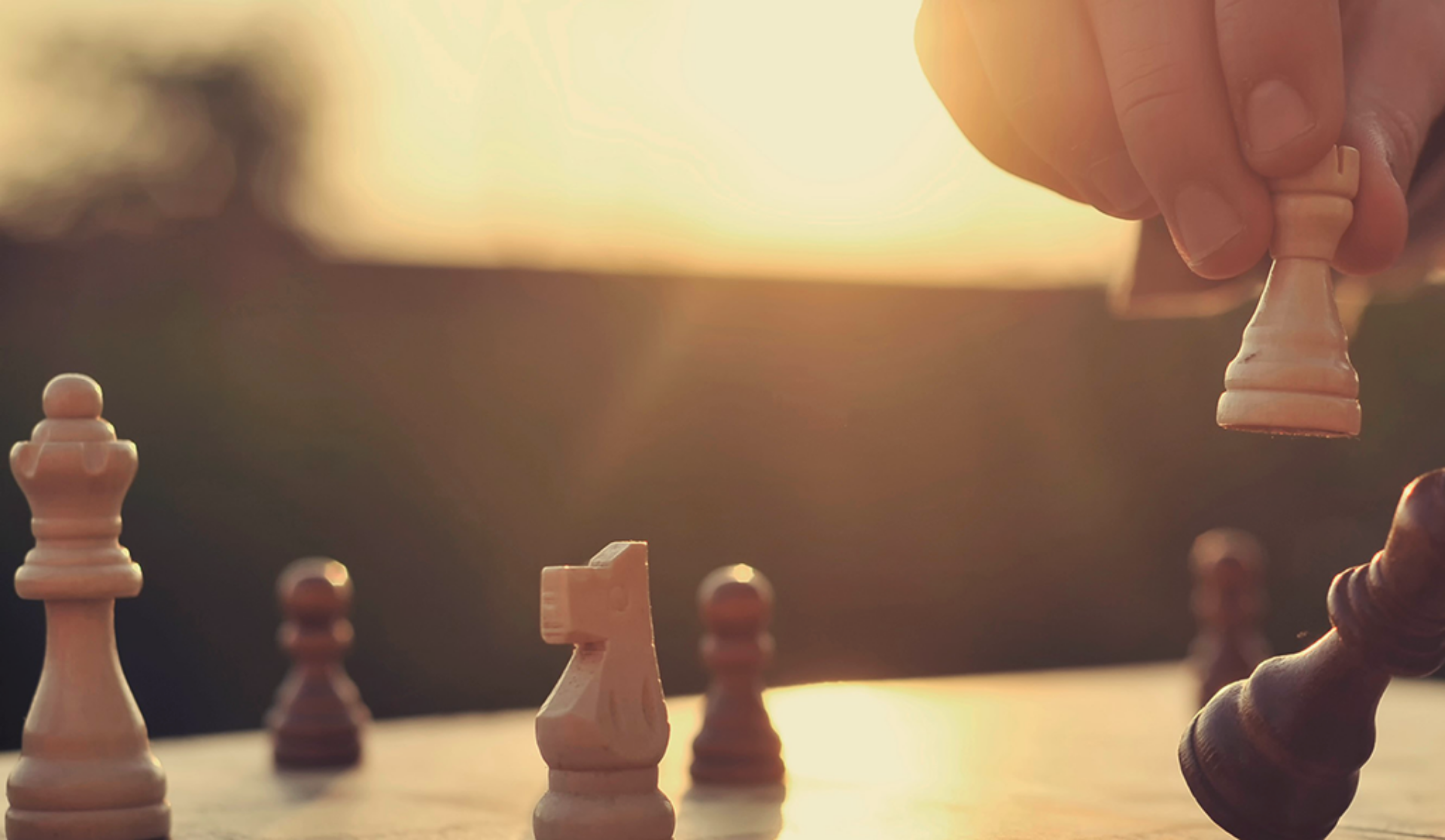 An individual playing chess, strategically positioning wooden pieces on a traditional chessboard, against the backdrop of a warm sunset.