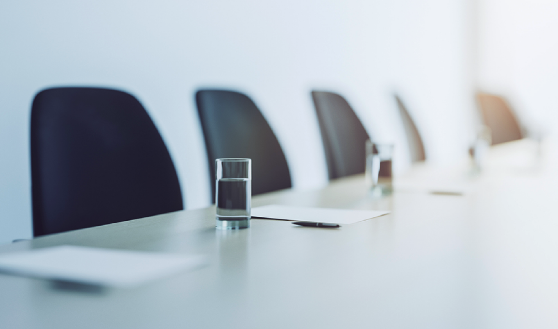 A conference table with black chairs, water glasses, notepads, and pens arranged neatly. The scene is brightly lit with a minimalistic setting.